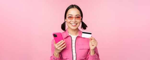 Compras en línea Compradora asiática sonriente con teléfono inteligente y pago con tarjeta de crédito en la aplicación móvil de pie sobre fondo rosa