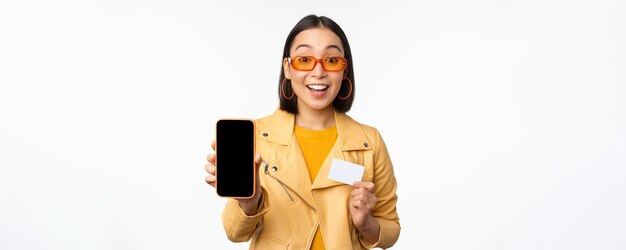 Compras en línea y concepto de personas Elegante mujer asiática que muestra la pantalla del teléfono móvil y la aplicación del teléfono inteligente con tarjeta de crédito de pie sobre fondo blanco
