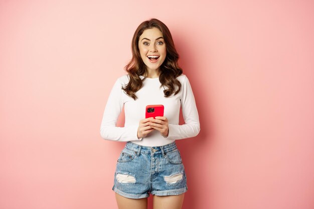 Compras en línea y concepto de teléfono móvil. Una chica atractiva emocionada sosteniendo un teléfono móvil y sonriendo, de pie sobre un fondo rosa.