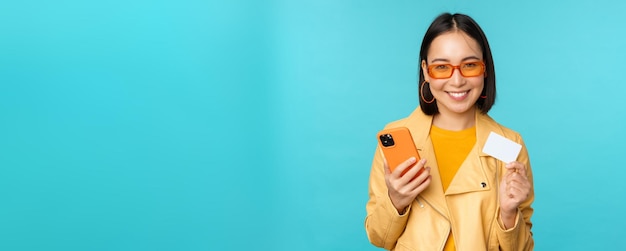 Compras en línea Elegante joven asiática con gafas de sol mostrando tarjeta de crédito y usando un teléfono inteligente pagando en internet haciendo una compra de pie sobre fondo azul