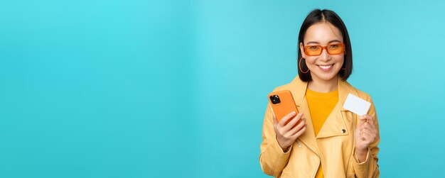 Compras en línea Elegante joven asiática con gafas de sol mostrando tarjeta de crédito y usando un teléfono inteligente pagando en internet haciendo una compra de pie sobre fondo azul