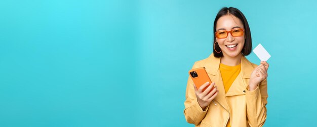 Compras en línea Elegante joven asiática con gafas de sol mostrando tarjeta de crédito y usando un teléfono inteligente pagando en internet haciendo una compra de pie sobre fondo azul