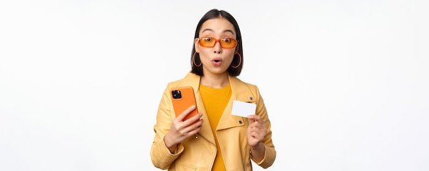 Compras en línea Elegante modelo femenino asiático en gafas de sol con tarjeta de crédito y teléfono móvil sonriendo feliz de pie sobre fondo blanco.
