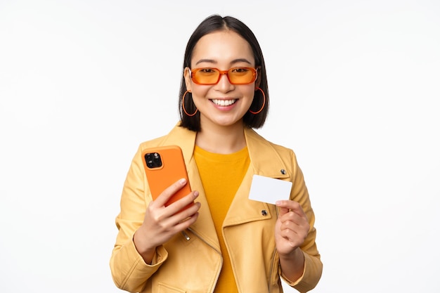 Compras en línea Elegante modelo femenino asiático en gafas de sol con tarjeta de crédito y teléfono móvil sonriendo feliz de pie sobre fondo blanco.