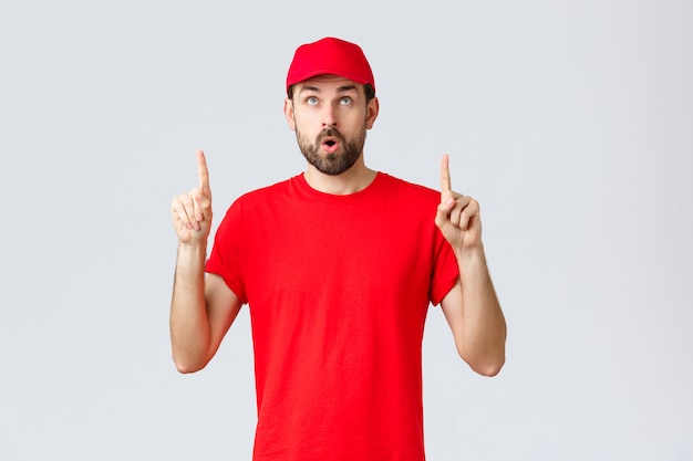 Compras en línea, entrega durante la cuarentena y concepto de comida para llevar. Empleado curioso e impresionado con gorra y camiseta de uniforme rojo, boca abierta interesada, leer pancarta, señalar con el dedo hacia arriba