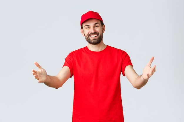 Compras en línea, entrega durante la cuarentena y concepto de comida para llevar. Mensajero agradable y amistoso en camiseta roja y gorra, uniforme de la empresa, extendiendo las manos hacia el cliente o el pedido, tomando el paquete