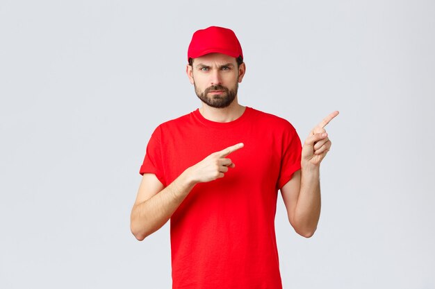 Compras en línea, entrega durante la cuarentena y concepto de comida para llevar. Mensajero enojado disgustado con gorra y camiseta de uniforme rojo, frunciendo el ceño malhumorado, señalando con el dedo en señal de desaprobación, se siente molesto