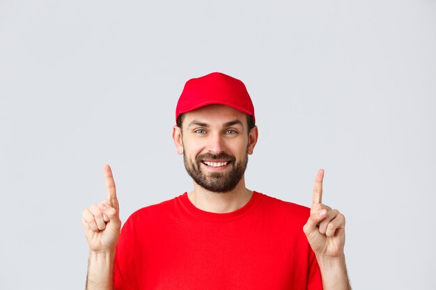 Compras en línea, entrega durante la cuarentena y concepto de comida para llevar. Mensajero sonriente barbudo alegre con gorra de uniforme rojo y camiseta, invitar a echar un vistazo a la promoción, apuntando con el dedo hacia arriba, fondo gris