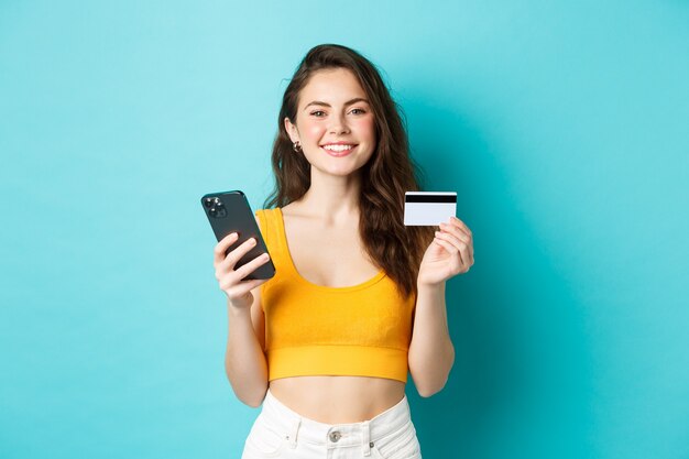 Las compras en línea. Hermosa mujer preparándose para las vacaciones de verano, reserva de boletos con tarjeta de crédito y aplicación para teléfono inteligente, de pie sobre fondo azul.