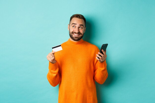 Las compras en línea. Hombre guapo pensando, sosteniendo el teléfono inteligente con tarjeta de crédito, pagando en la tienda de Internet, parado sobre una pared turquesa clara.