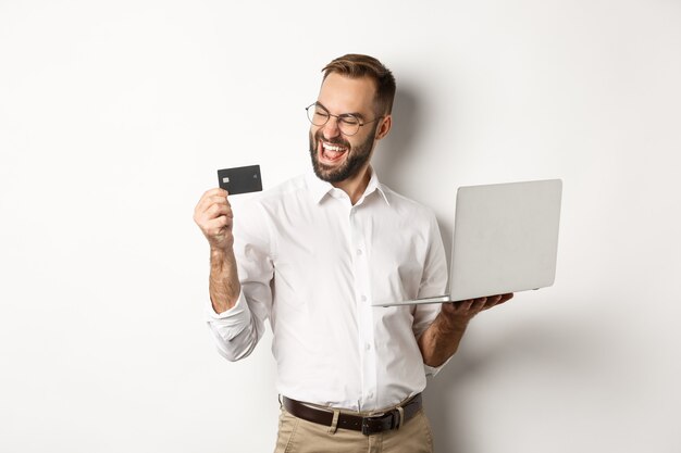 Las compras en línea. Hombre guapo satisfecho mirando la tarjeta de crédito después de hacer un pedido por internet, usando la computadora portátil, de pie