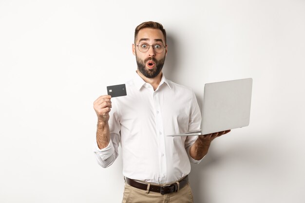 Las compras en línea. Hombre sorprendido que sostiene la computadora portátil y la tarjeta de crédito, tienda de internet de la tienda, de pie