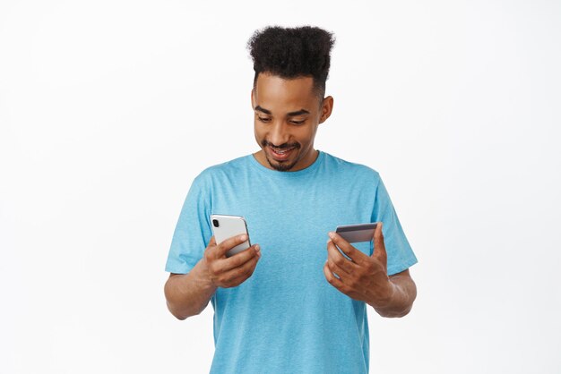 Compras en línea Joven afroamericano pagando en internet usando un teléfono inteligente y una tarjeta de crédito para hacer una compra de pie en una camiseta azul contra un fondo blanco