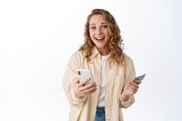 Compras en línea Joven chica positiva sonriendo usando un teléfono inteligente y pagando con tarjeta de crédito de plástico de pie en ropa elegante contra fondo blanco