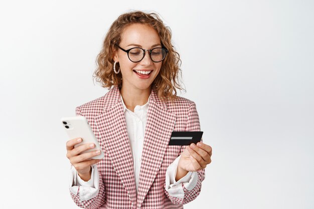 Las compras en línea. Joven empresaria pagando en la aplicación de teléfono inteligente, sosteniendo el teléfono móvil y la tarjeta de crédito, de pie en blanco.