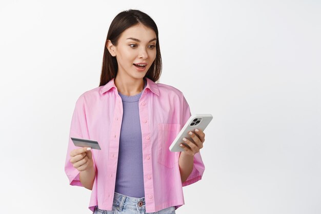 Compras en línea Mujer joven emocionada con tarjeta de crédito que parece sorprendida por el pedido de la aplicación de teléfono móvil en la aplicación de pie sobre fondo blanco