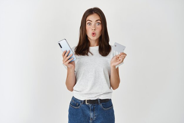 Las compras en línea. Mujer joven morena haciendo compras en internet con tarjeta de crédito y smartphone, mirando al frente divertido, pared blanca.
