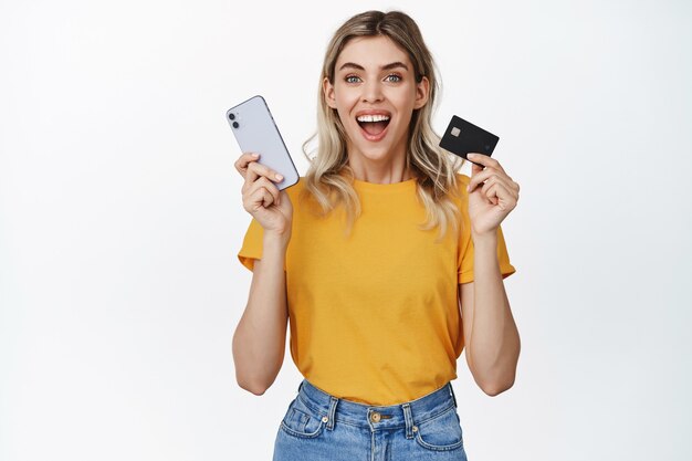 Foto gratuita las compras en línea. mujer joven con teléfono móvil y tarjeta de crédito mirando emocionado, comprando en la aplicación, pagando algo, haciendo una compra en un teléfono inteligente en blanco