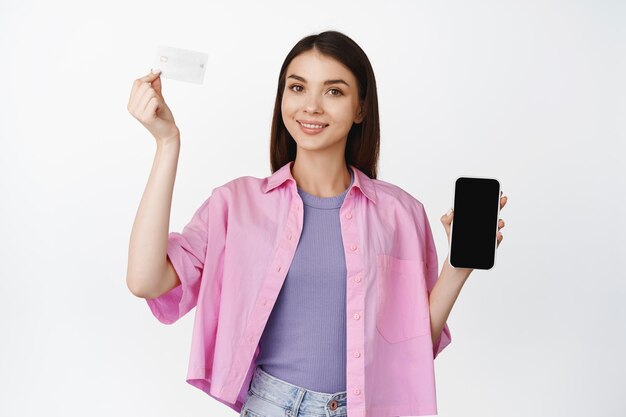 Compras en línea Mujer morena sonriente levantando la mano con tarjeta de crédito que muestra la interfaz móvil de la pantalla del teléfono inteligente de pie sobre fondo blanco