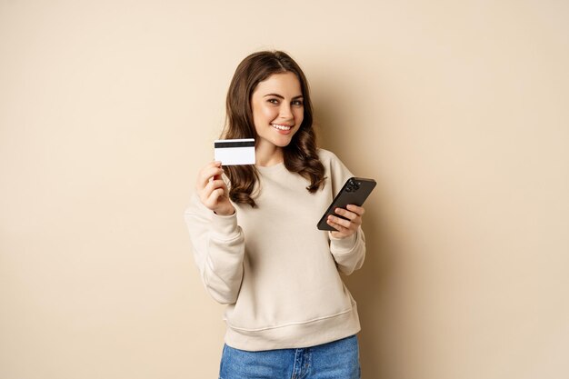 Las compras en línea. Mujer morena sonriente mostrando tarjeta de crédito, usando una aplicación de teléfono inteligente, de pie sobre un fondo beige.