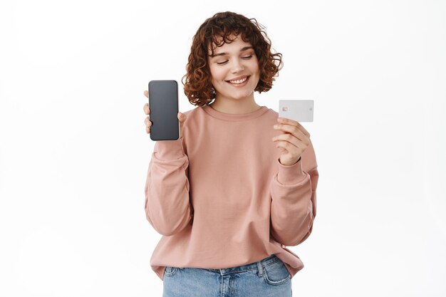 Las compras en línea. Mujer sonriente que muestra la pantalla en blanco del teléfono inteligente, la aplicación móvil y se ve satisfecha con la tarjeta de crédito bancaria, paga algo, hace un pedido, se para sobre fondo blanco