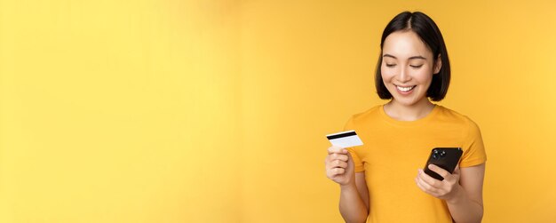 Compras en línea Niña asiática sonriente usando tarjeta de crédito y aplicación de teléfono móvil pagando un pedido sin contacto en la aplicación de teléfono inteligente de pie sobre fondo amarillo