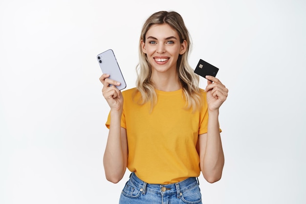 Compras en línea Niña rubia sonriente que muestra la tarjeta de crédito y sostiene el teléfono móvil pagando en la tienda de Internet usando la aplicación de dinero de pie sobre fondo blanco