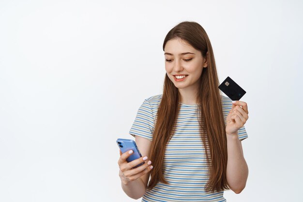 Las compras en línea. Niña sonriente con tarjeta de crédito y teléfono inteligente, pagando con una aplicación de banca móvil, comprando en una tienda de Internet, fondo blanco