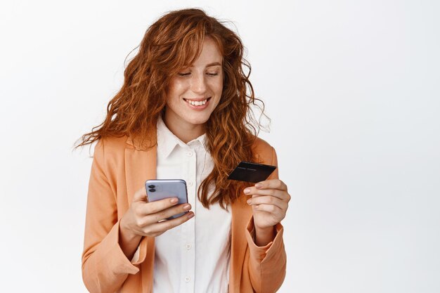 Compras en línea Orden de empresaria pelirroja sonriente en el teléfono móvil con tarjeta de crédito y teléfono inteligente de pie en traje contra el fondo blanco