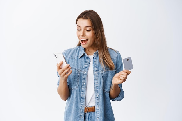 Las compras en línea. Retrato de mujer joven emocionada leyendo la pantalla del teléfono inteligente y sosteniendo una tarjeta de crédito de plástico, pagando la compra por Internet, haciendo un pedido, blanco