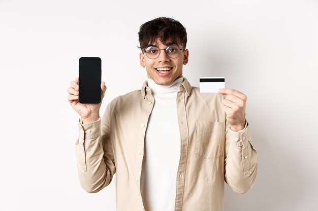 Compras en línea sorprendido y feliz joven mostrando tarjeta de crédito y pantalla de teléfono móvil de pie ...