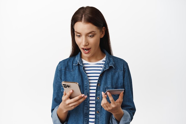 Foto gratuita compras. mujer adulta joven pagando por teléfono móvil, con tarjeta de crédito y mirando el teléfono inteligente con cara emocionada, venta especial en la tienda de internet, fondo blanco