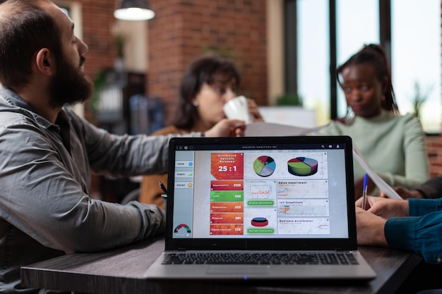 Computadora portátil con gráficos de la empresa en la pantalla de pie sobre la mesa en la oficina de inicio mientras diversos equipos comparten documentos con el volumen de negocios de marketing en él. Empresarios multiétnicos analizando gráficos