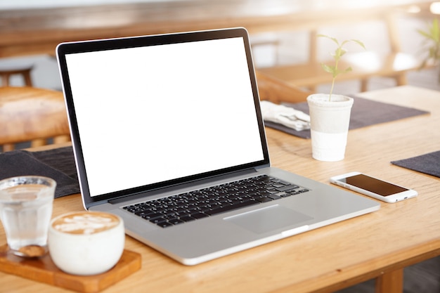 computadora portátil con pantalla en blanco. Espacio de trabajo del joven autónomo: abrir un ordenador portátil, un teléfono móvil y un capuchino en la mesa de madera