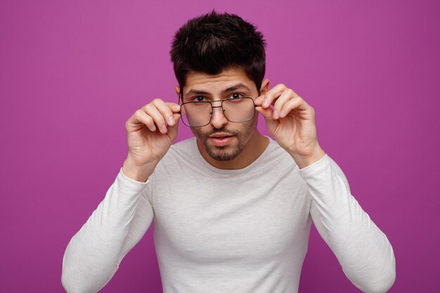 Concentrado joven apuesto hombre con gafas mirando a la cámara desde detrás de las gafas mientras mantiene las manos sobre ellos sobre fondo púrpura