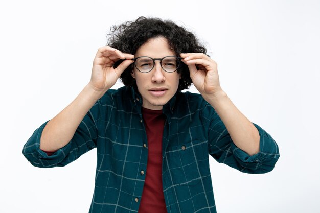 Concentrado joven apuesto hombre con gafas mirando a la cámara mientras agarra gafas aislado sobre fondo blanco.