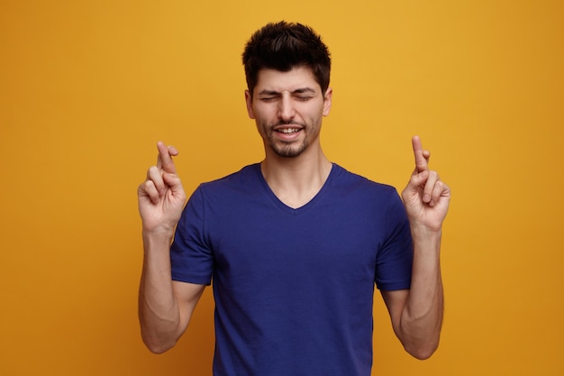 Concentrado joven guapo cruzando los dedos pidiendo deseo con los ojos cerrados sobre fondo amarillo