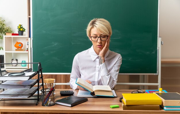 Concentrado joven maestra rubia con gafas sentado en un escritorio con útiles escolares en el aula leyendo el libro manteniendo las manos en la cara