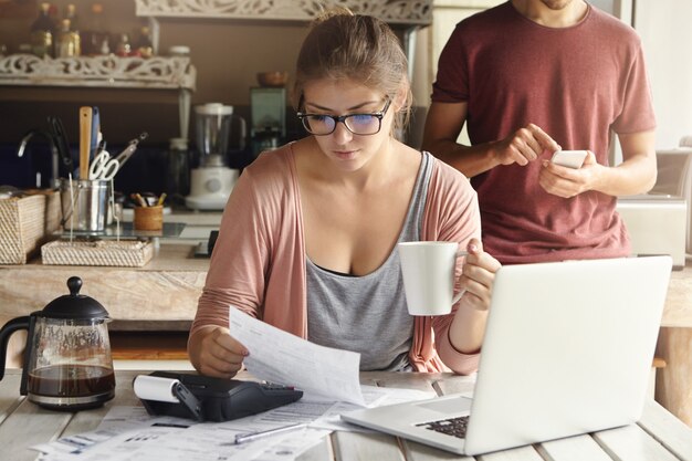 Concentrado joven mujer caucásica tomando café por la mañana mientras trabaja con las finanzas en la cocina