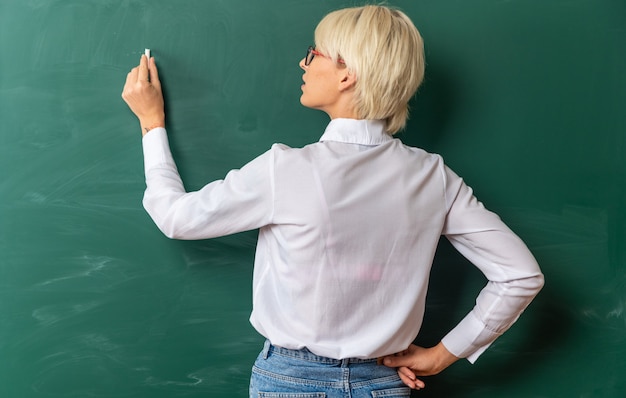 Foto gratuita concentrado joven profesora rubia con gafas en el aula de pie detrás de la vista delante de la pizarra escribiendo en la pizarra con tiza manteniendo la mano en la cintura