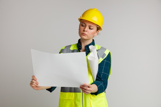 Concentrado joven trabajador de la construcción con casco de seguridad y chaleco de seguridad sosteniendo papeles mirando uno