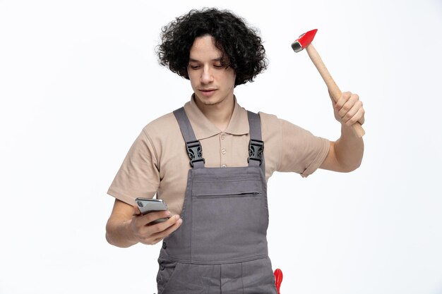 Concentrado joven trabajador de la construcción masculino vistiendo uniforme levantando el martillo mientras sostiene y mira el teléfono móvil con un destornillador en el bolsillo aislado sobre fondo blanco.