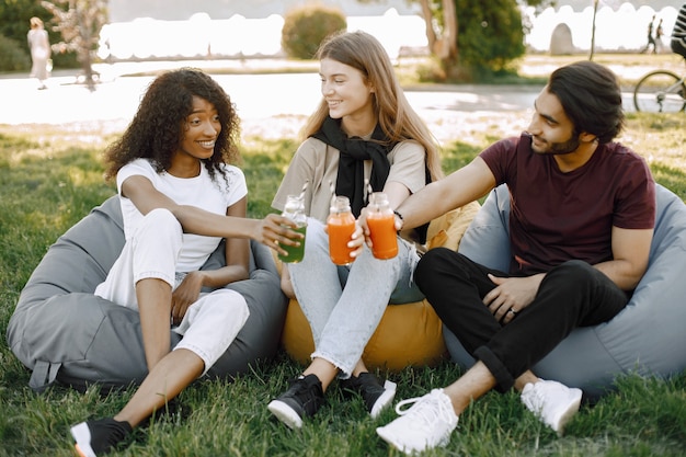 Concéntrese en botellas. Amigos colgando en el parque verde, relajándose, sentados en los sillones puf sobre el césped