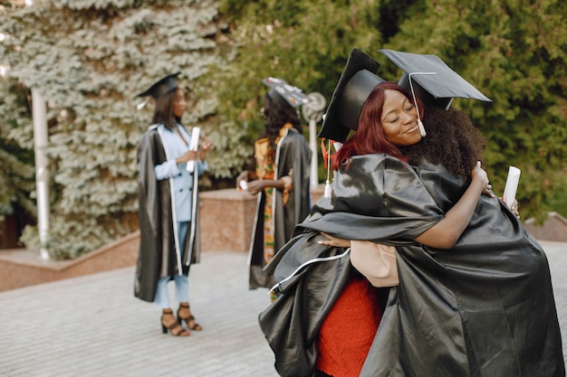 Concéntrese en dos jóvenes estudiantes afroamericanas vestidas con un traje de graduación negro. Campus como telón de fondo. Niñas, abrazar