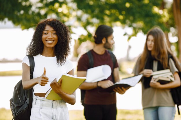 Concéntrese en una niña africana que está de pie por separado y muestra el pulgar hacia arriba. Grupo de estudiantes internacionales parados juntos en el parque en la universidad