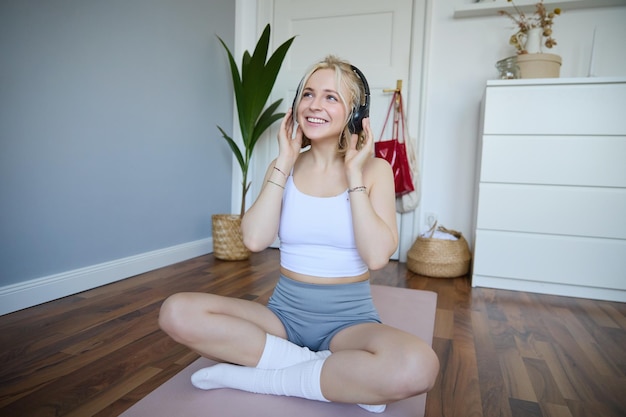 Foto gratuita concept de deporte y bienestar mujer sonriente con auriculares inalámbricos sentada en una alfombra de yoga escuchando