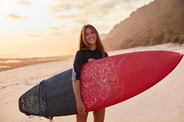 Concepto de actividades de verano. Satisfecha hermosa joven vestida con traje de baño, lleva tabla larga, tiene vacaciones en el extranjero en el país turístico