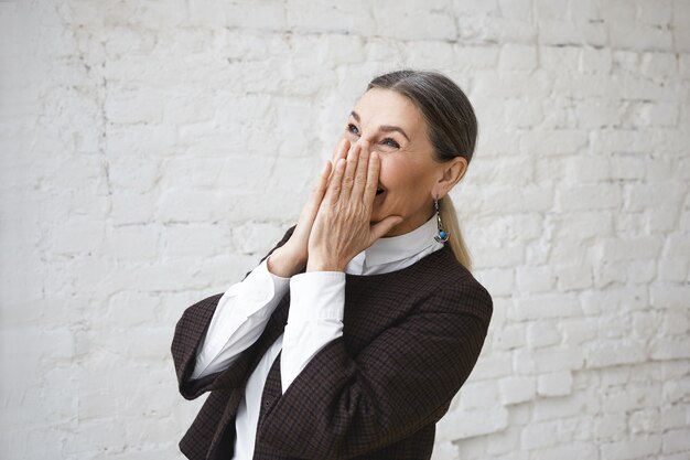 Concepto de alegría, diversión, emociones humanas positivas y sentimientos. Retrato de feliz alegre mujer madura vistiendo camisa blanca y chaqueta que cubre la boca mientras se ríe de la broma o se regocija con las buenas noticias