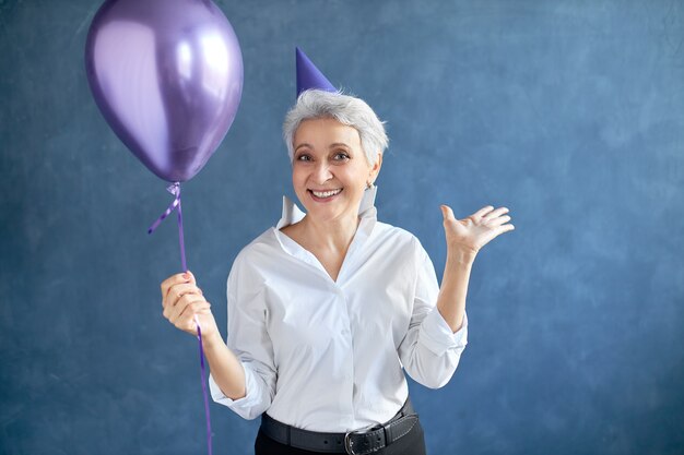 Concepto de alegría, felicidad, diversión y emociones positivas. Retrato de una hermosa mujer de pelo gris de 50 años emocionada con sombrero cónico en la cabeza