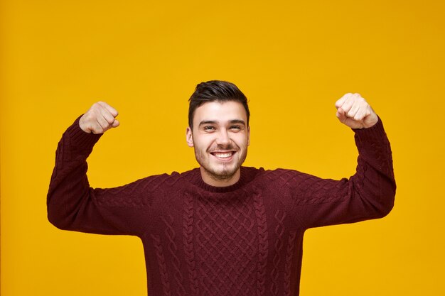 Concepto de alegría, felicidad, ganar y éxito. Feliz carismático joven varón de raza caucásica en elegante suéter apretando los puños, expresando una reacción genuina ante las buenas noticias, con mirada emocionada